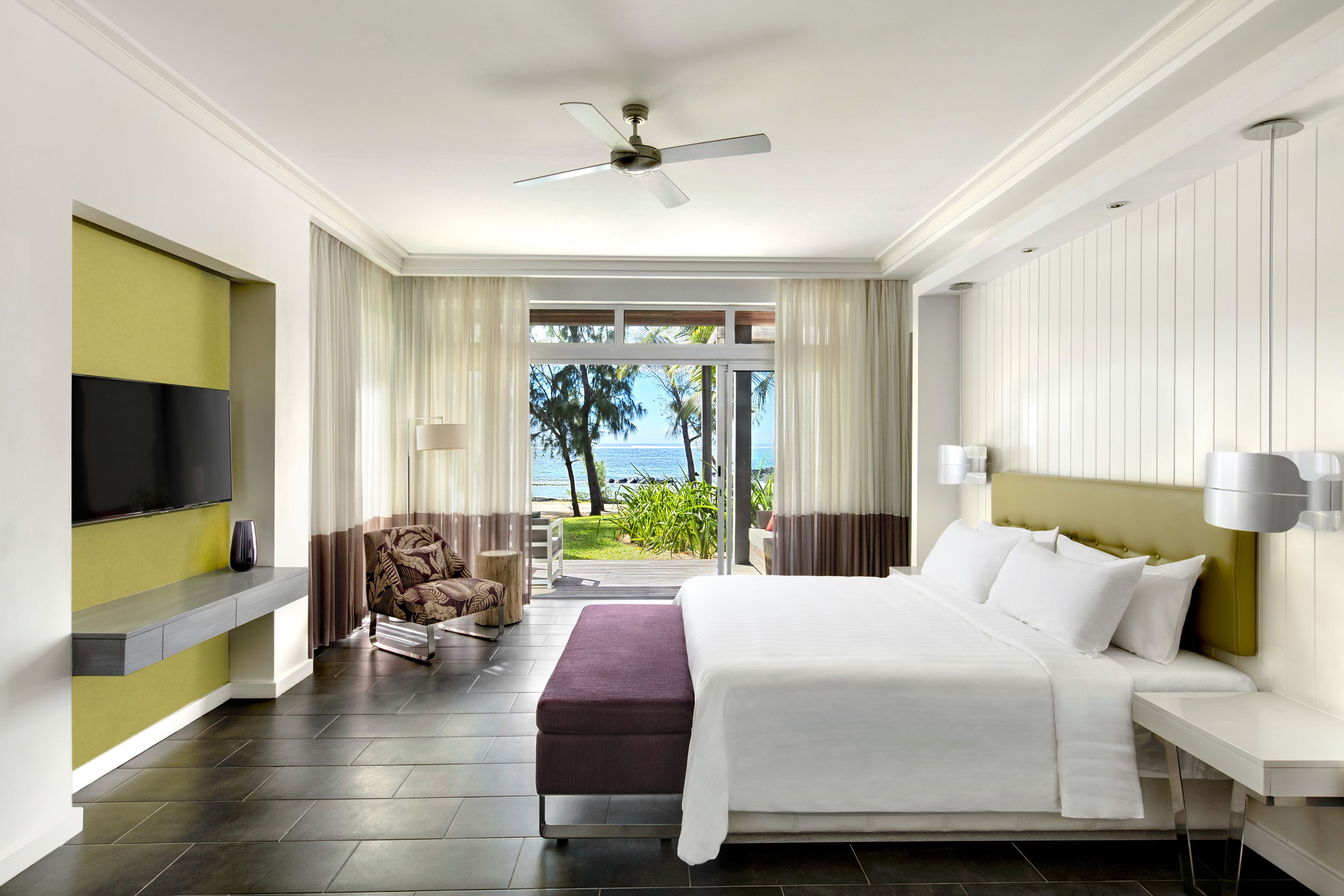 A modern hotel room with a large bed, a television, a seating area with a chair, and a view of the ocean.