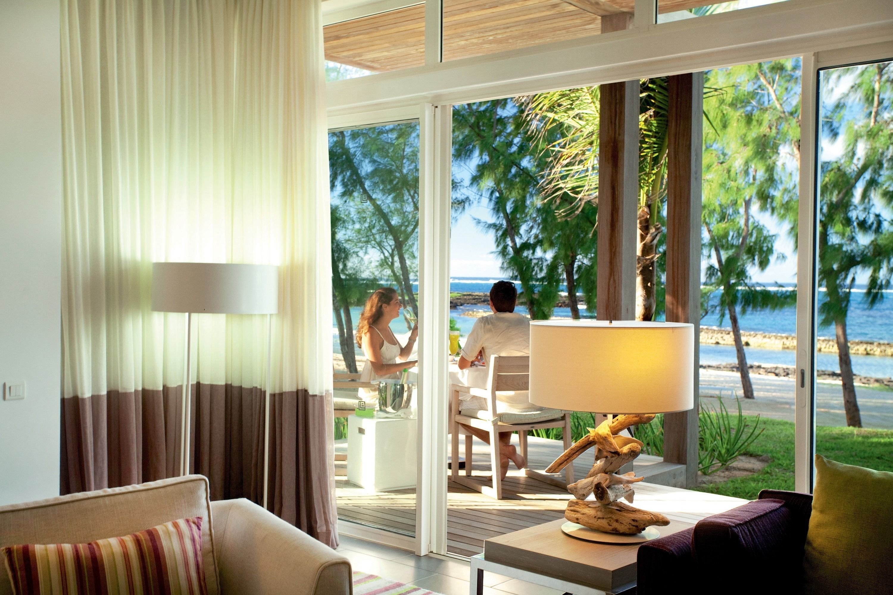 Una habitación de hotel moderna con un amplio ventanal corredero que da a un balcón con vistas al mar y palmeras.