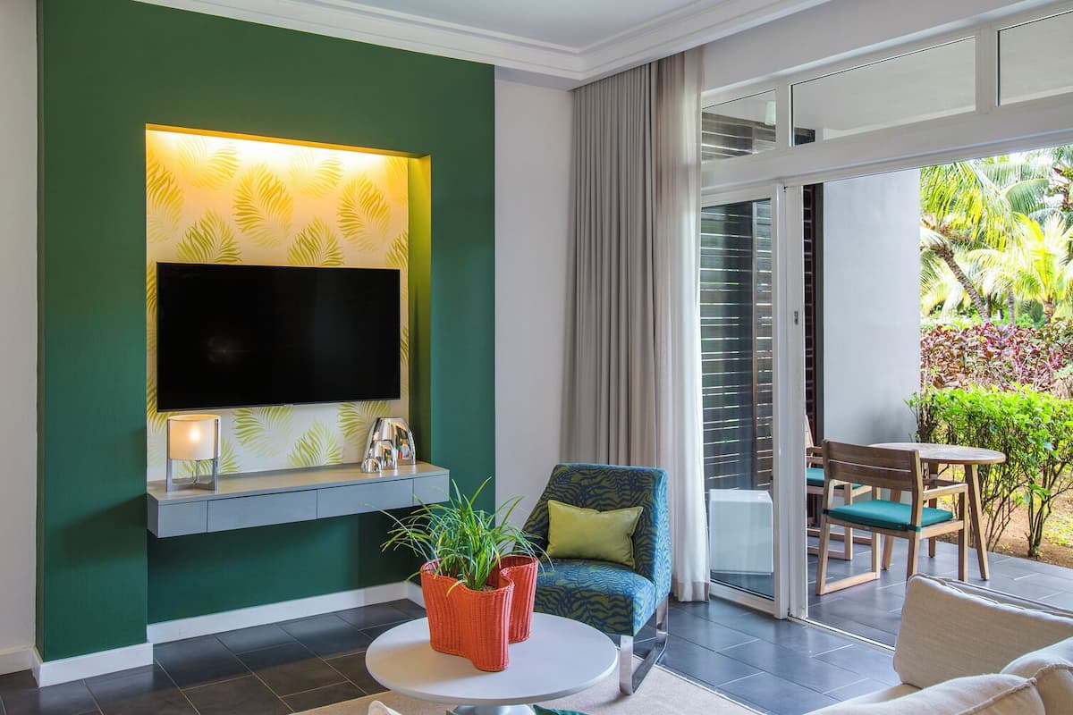 Un salon moderne avec un téléviseur à écran plat fixé sur un mur vert, un ventilateur de plafond, une table basse ronde agrémentée de plantes en pot, un canapé beige et une porte-fenêtre coulissante donnant sur un balcon avec des meubles d’extérieur.