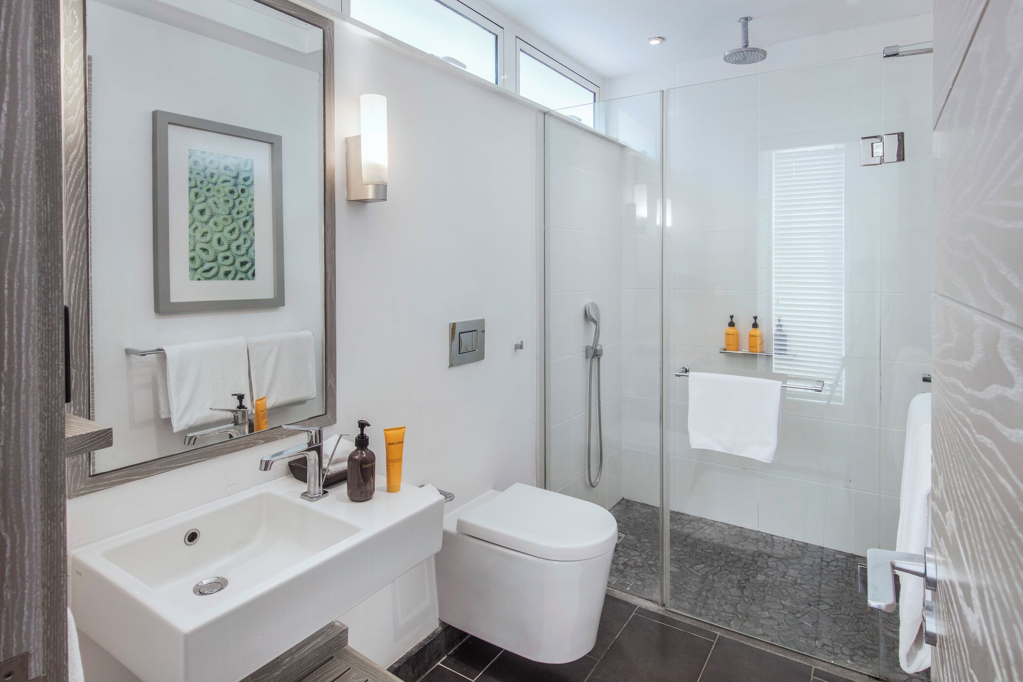 Un bagno moderno con lavandino, water e doccia bianchi. Sopra lo specchio, un'opera d'arte incorniciata e un porta asciugamani con un asciugamano.