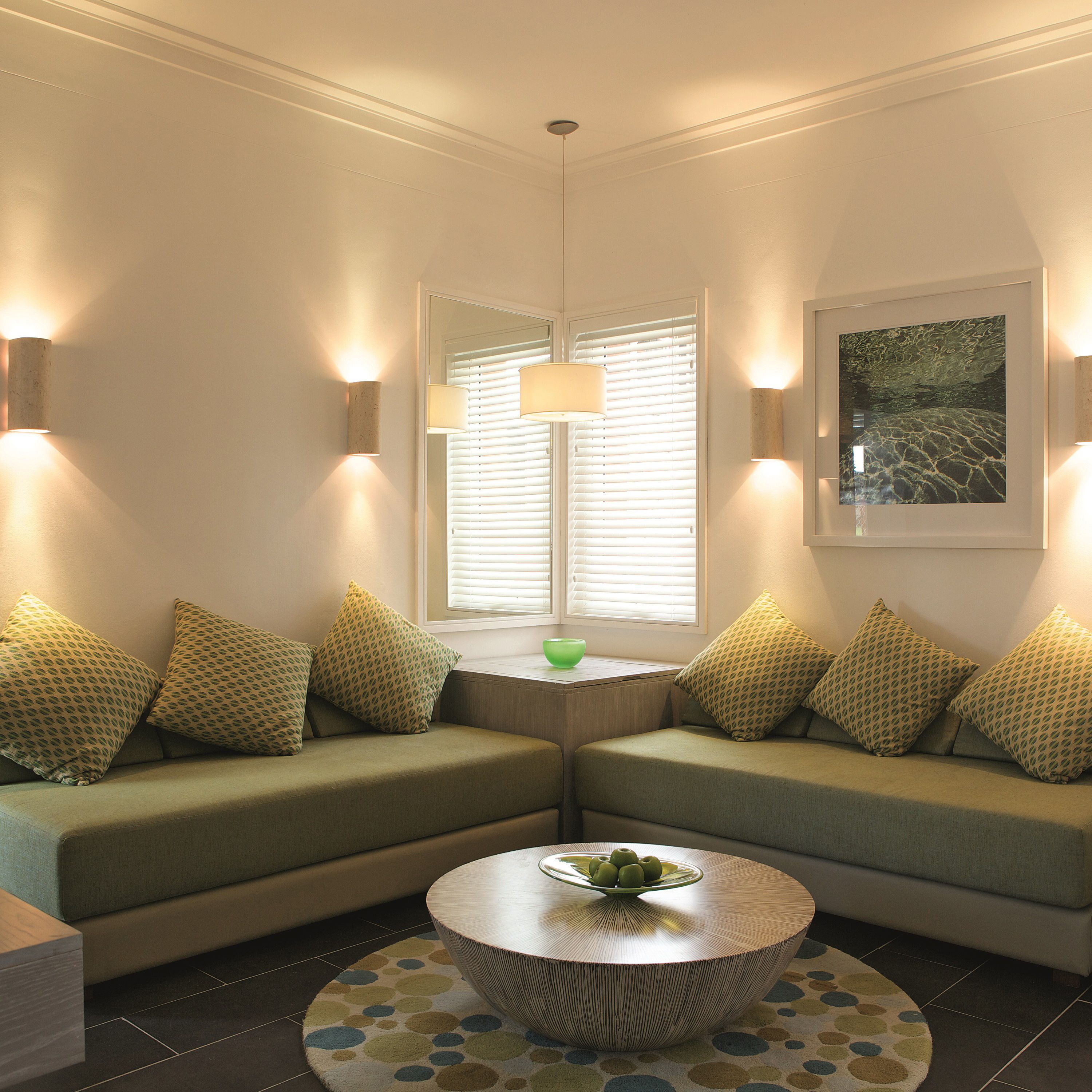 A modern living room with two green sofas, a round coffee table with fruit, and a green bowl on the windowsill.