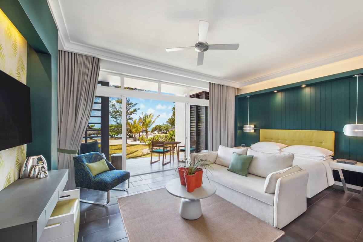 Une chambre d’hôtel moderne avec un canapé, un fauteuil, une table à manger, un lit avec une tête de lit jaune et un téléviseur fixé au mur.