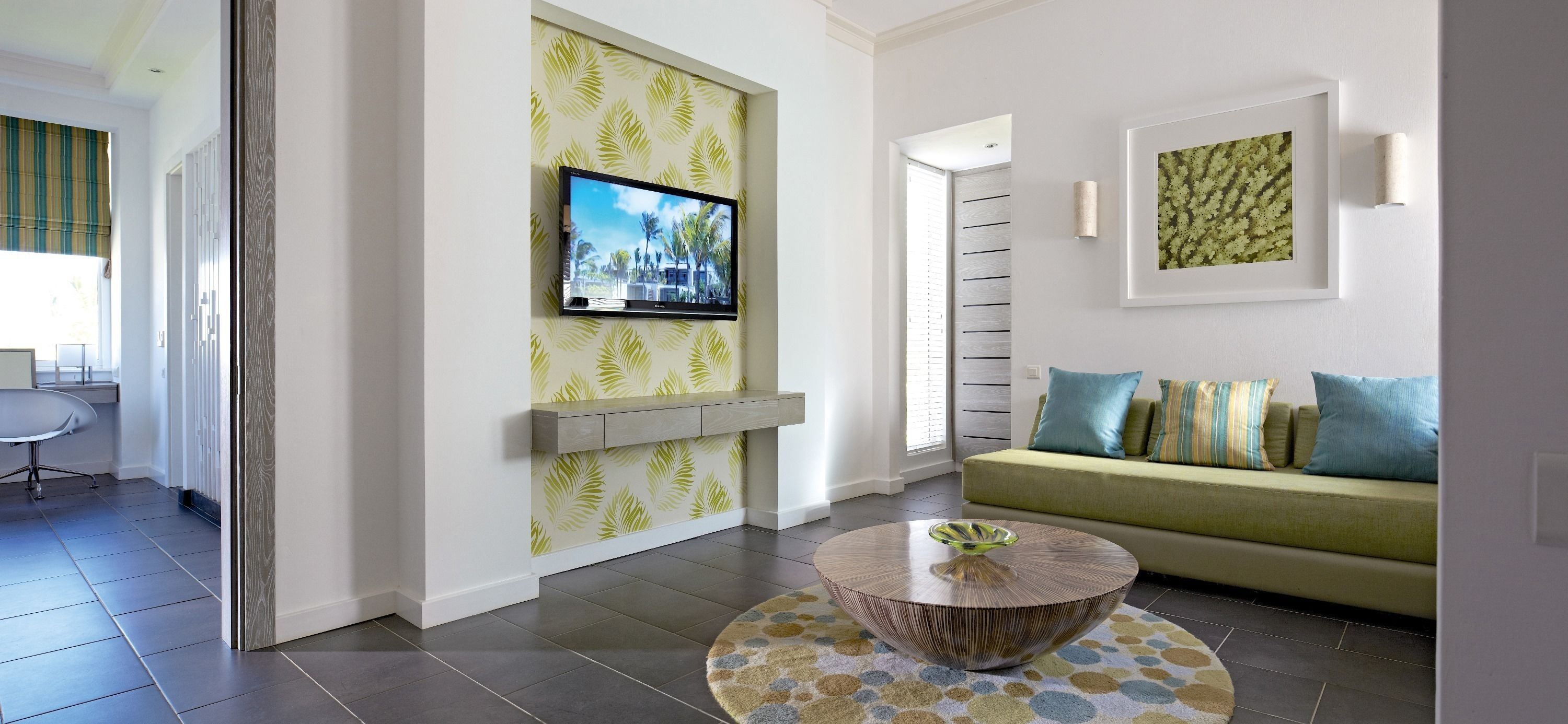 A modern living room with a flat-screen TV mounted on a wall with leaf-pattern wallpaper, a green sofa with patterned cushions, and a round coffee table with a decorative base.
