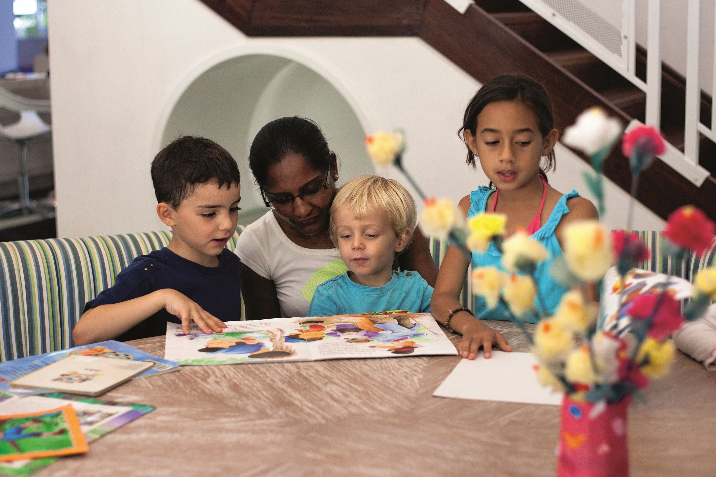Un groupe d’enfants et un adulte sont assis autour d’une table, devant des livres et un vase rempli de fleurs.