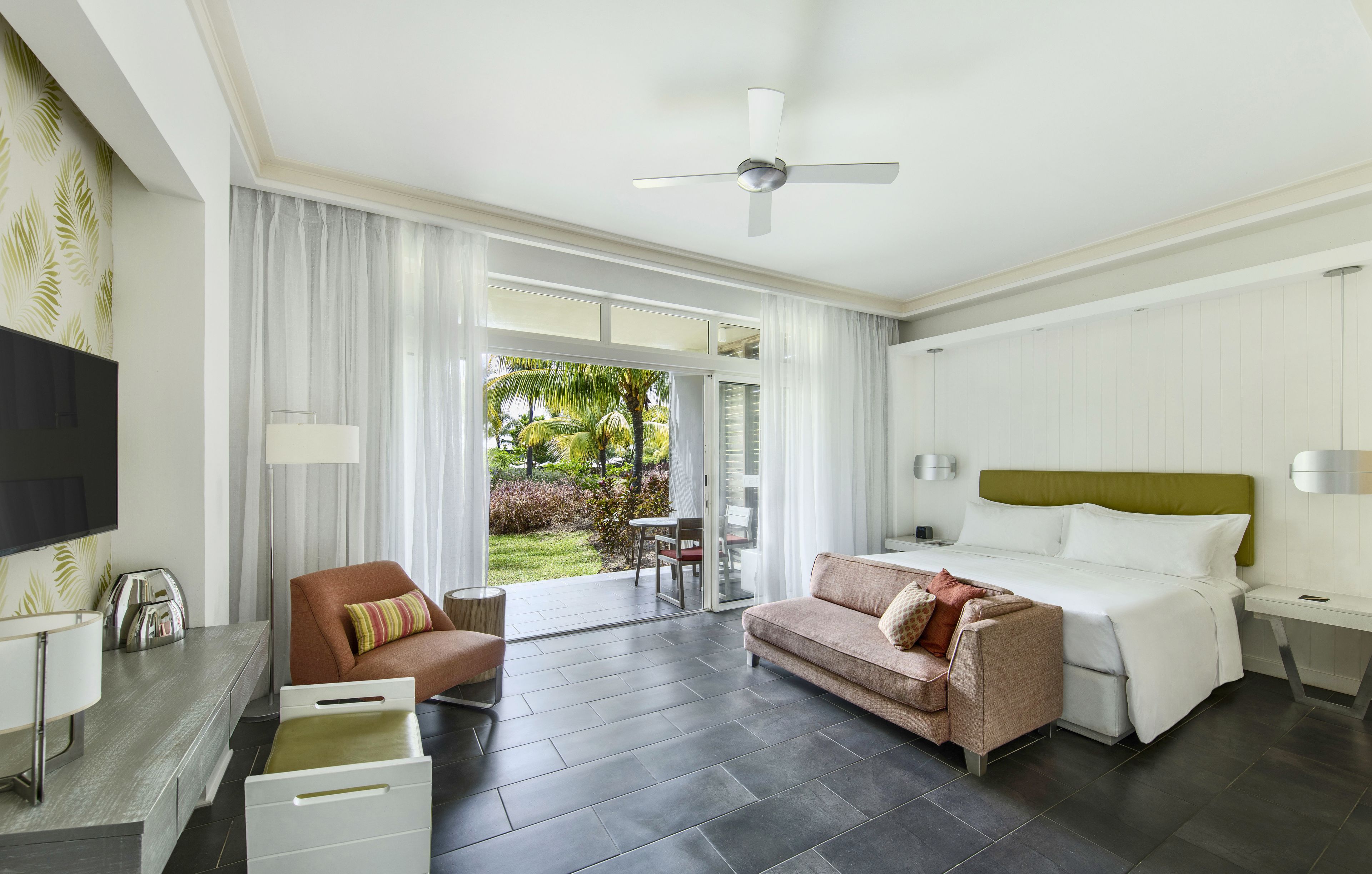 A hotel room with a bed, a sofa, a TV, a desk, and a balcony with a view of the garden.