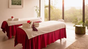 A room with two beds, each with a red bedspread and white linens, a large window with a view of greenery, and a potted plant on the windowsill.