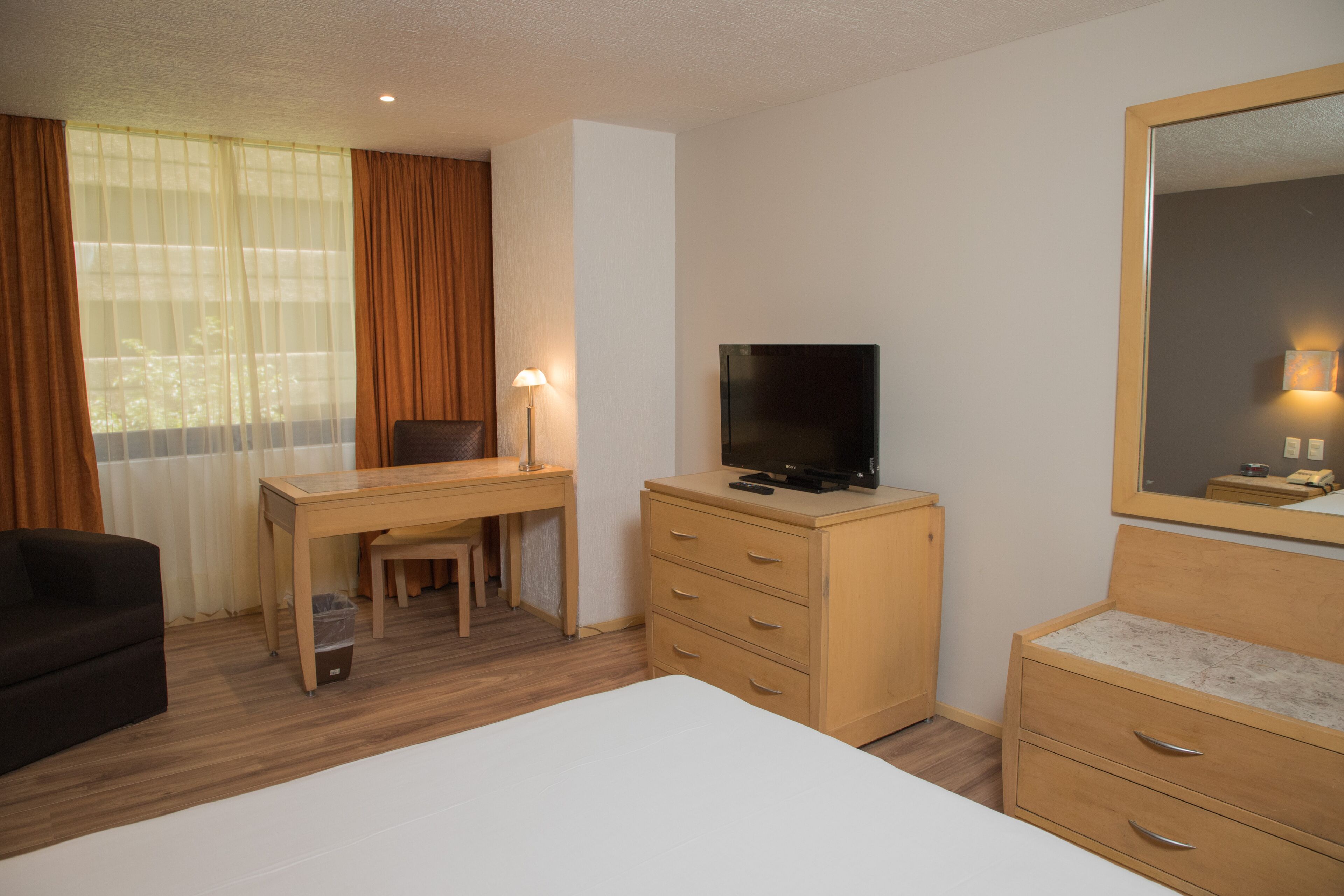 Habitación de hotel con cama, escritorio, silla, televisor, cómoda y ventana con cortinas.