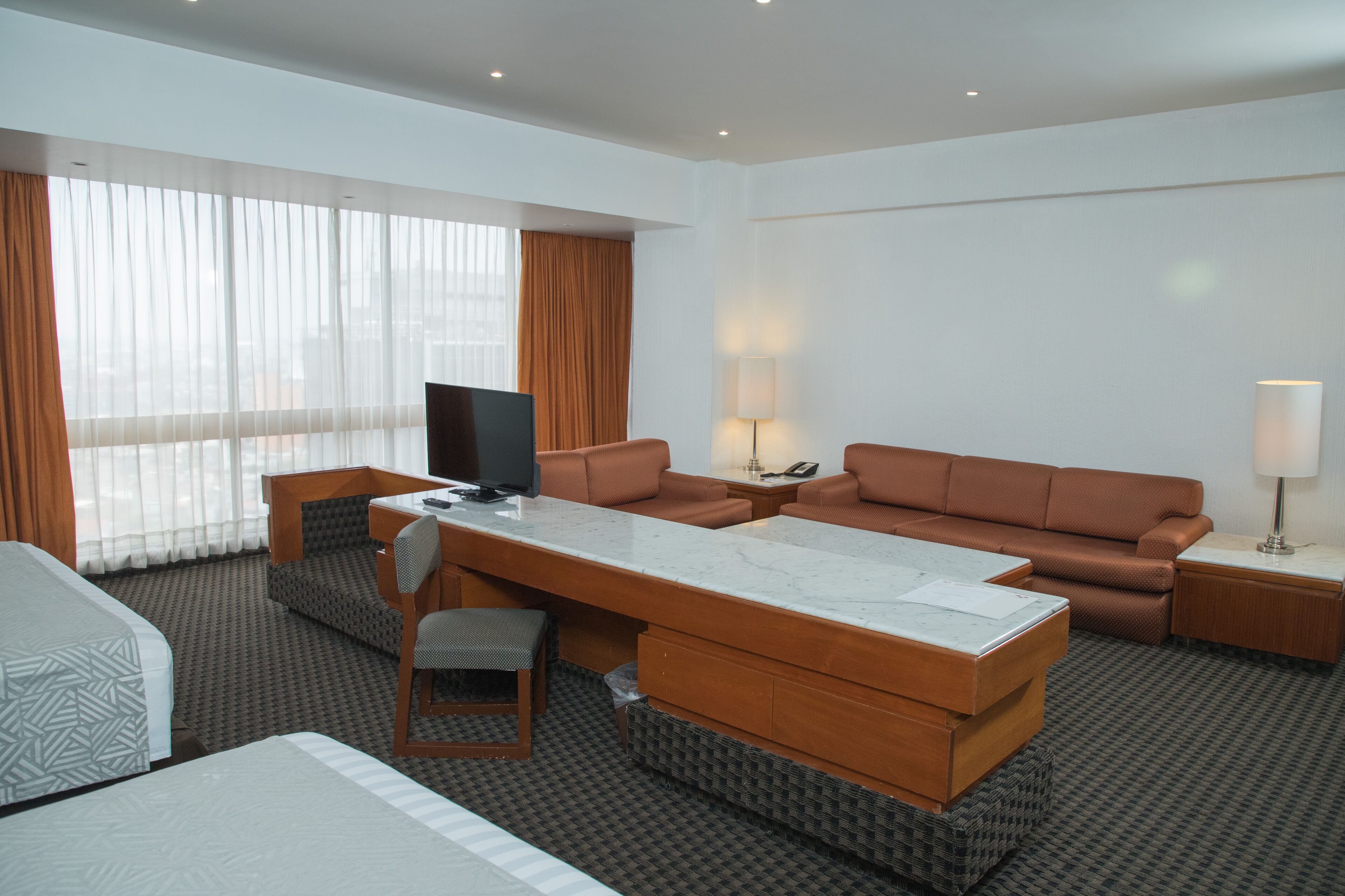 A hotel room with a bed, a sofa, a desk with a TV, and a large window with curtains.