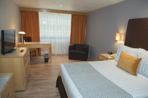 Una habitación de hotel con cama, escritorio, silla, televisor y mesita de noche con lámpara.