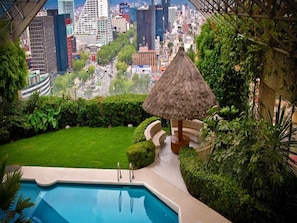 A pool area with a thatched umbrella, surrounded by lush greenery and a view of a cityscape. - Sevilla Palace Hotel (Mexico City)