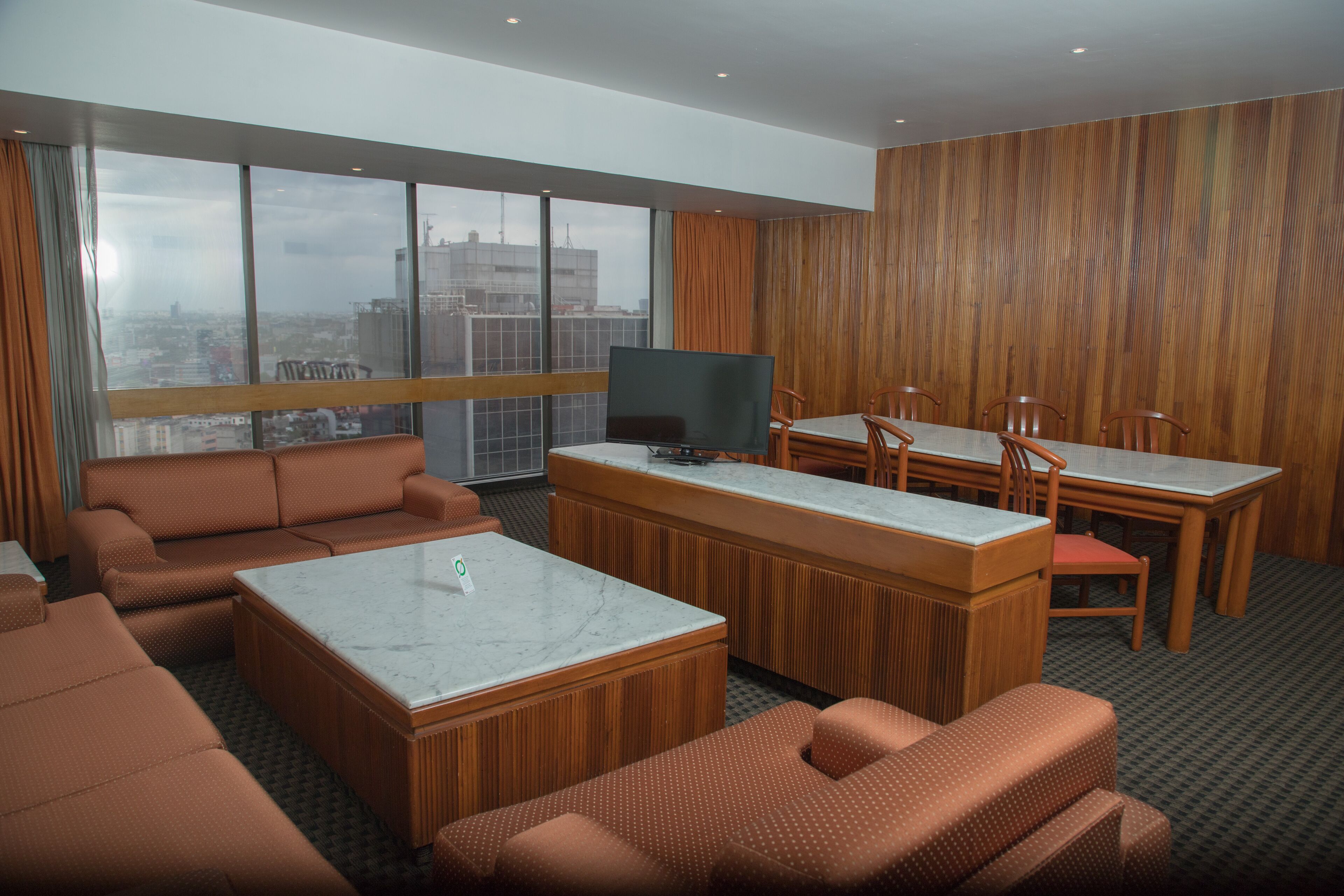 A modern hotel room with a large window, a sofa, a coffee table, a TV, and a dining area with chairs and a table.