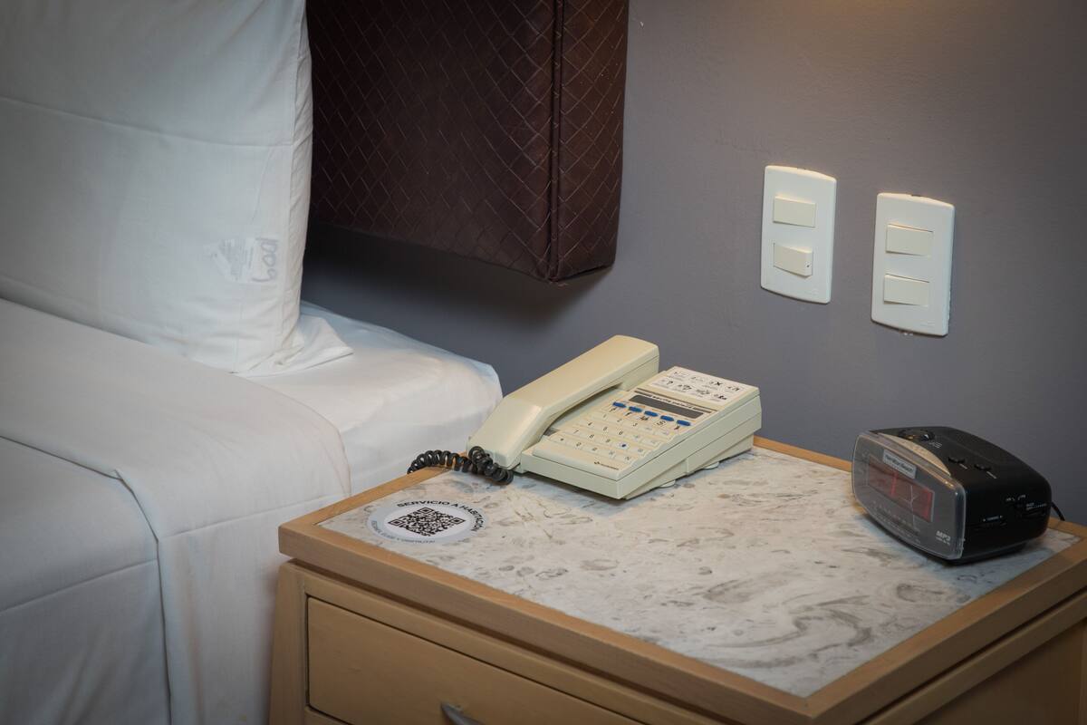 Hotellihuone, jossa on sänky, yöpöytä, jossa on puhelin ja radio, sekä kaksi seinään asennettua valokytkintä.