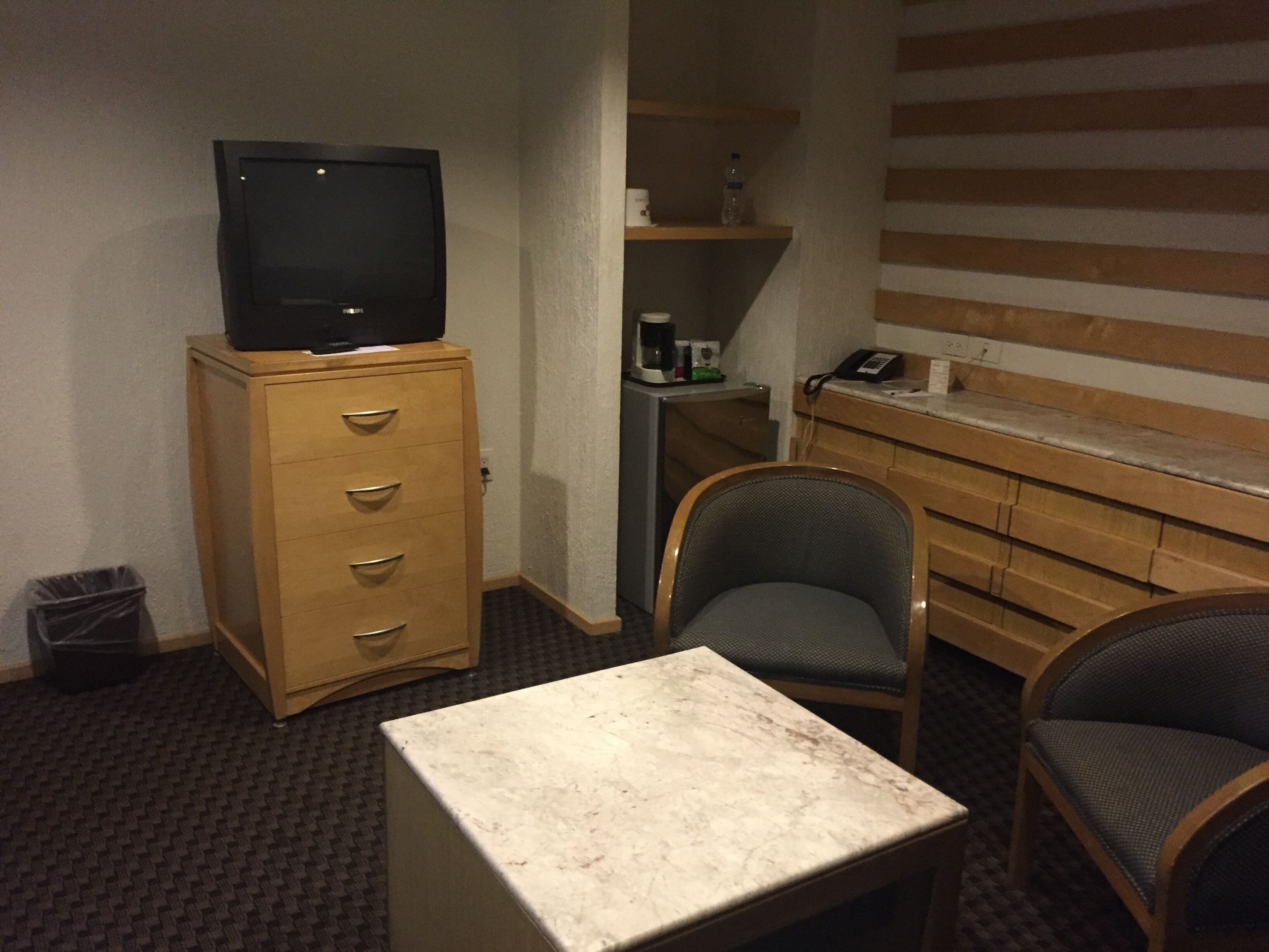 A hotel room with a wooden cabinet, a television on top, a small table, two chairs, a trash bin, and a minibar with a phone and a bottle.