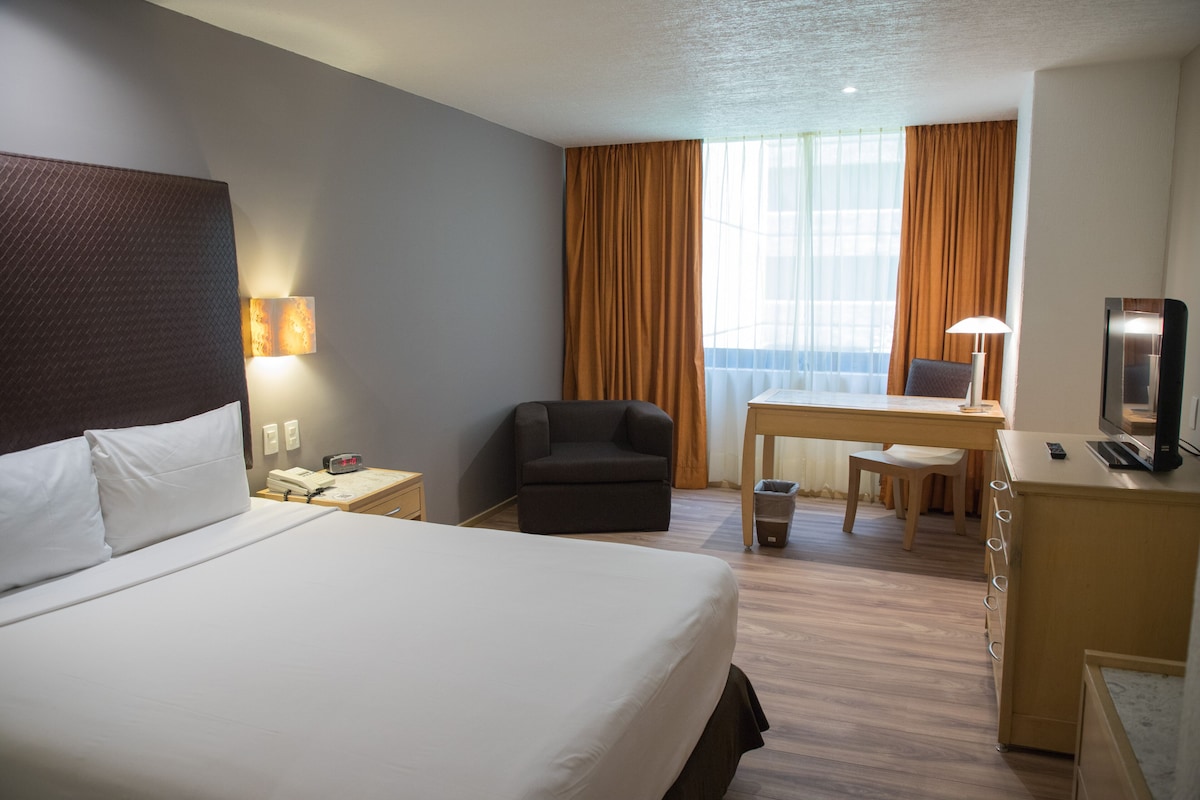 Habitación de hotel con una cama grande, un escritorio con una silla, una mesita, una lámpara y un televisor.