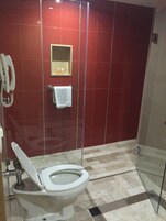 A bathroom with a toilet, a glass shower enclosure, a red tiled wall, and a phone mounted on the wall.