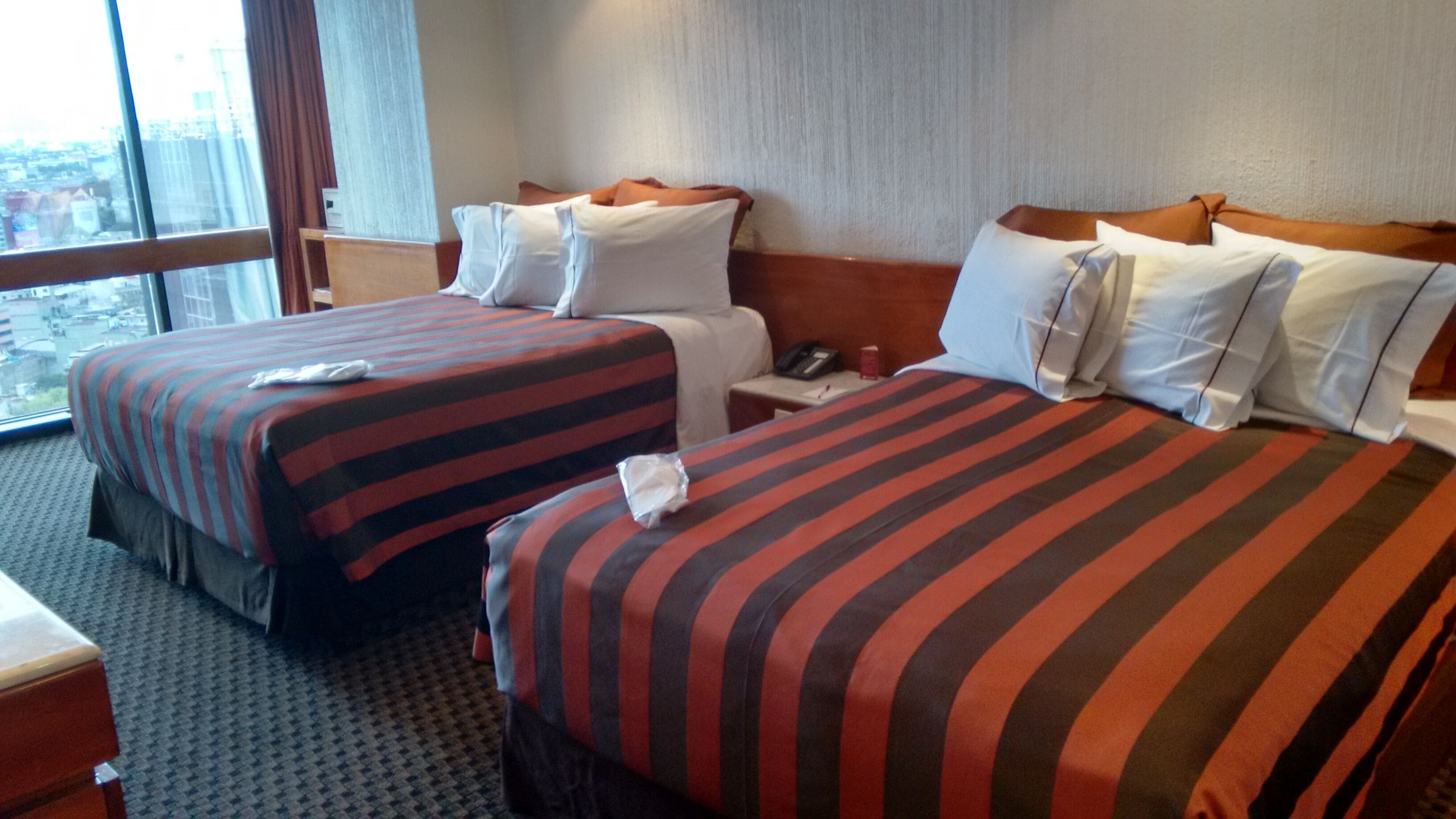 Habitación de hotel con dos camas, cada una con ropa de cama a rayas, una mesita de noche con teléfono y una ventana con vista a la ciudad.
