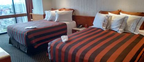 Habitación de hotel con dos camas, cada una con ropa de cama a rayas, una mesita de noche con teléfono y una ventana con vista a la ciudad.