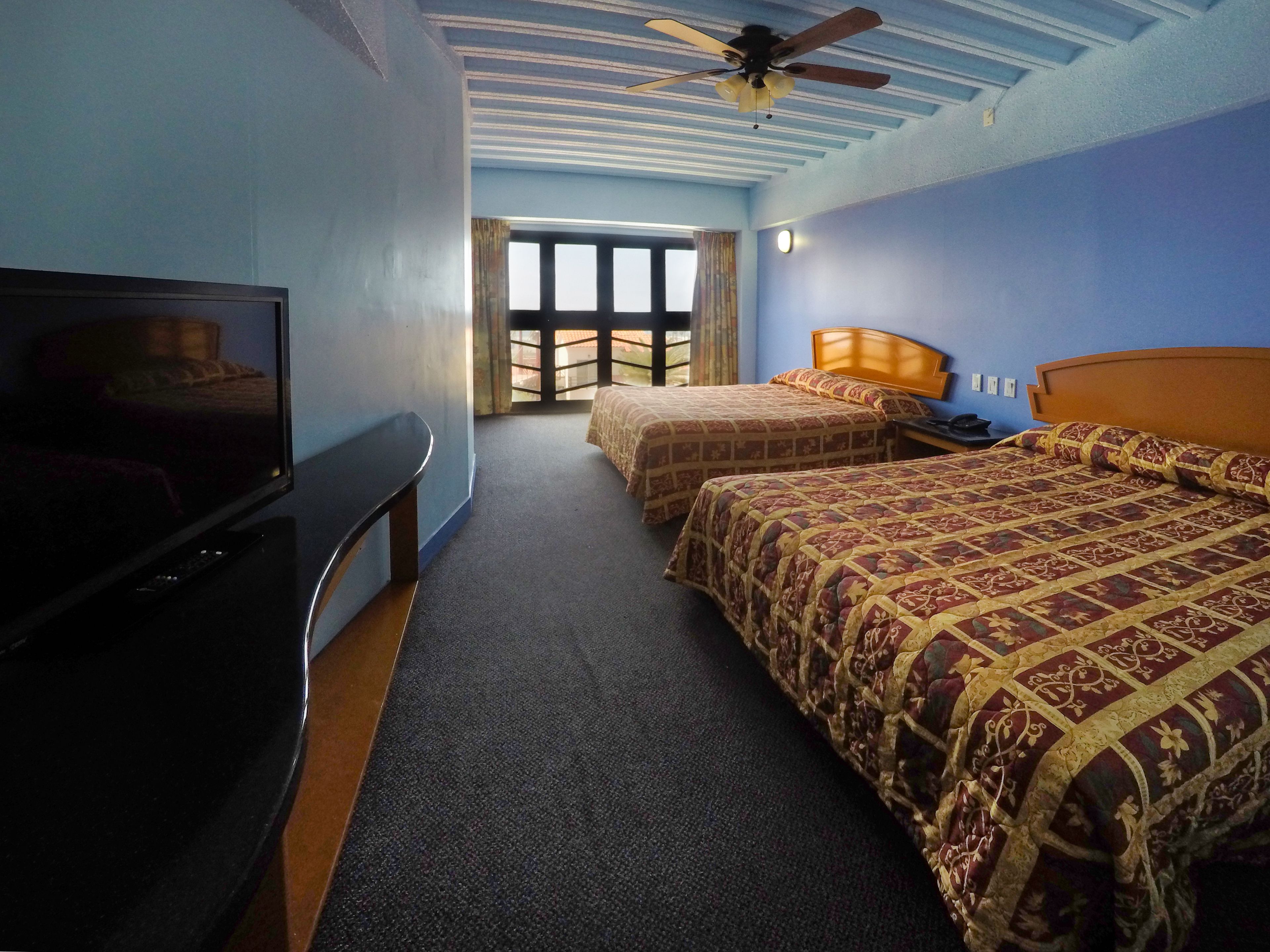 A hotel room with two beds, a television, a ceiling fan, and a large window.