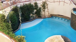 Ein Pool mit Natursteinrand, umgeben von viel Grün und einer Liegefläche mit Stühlen und einem Tisch.