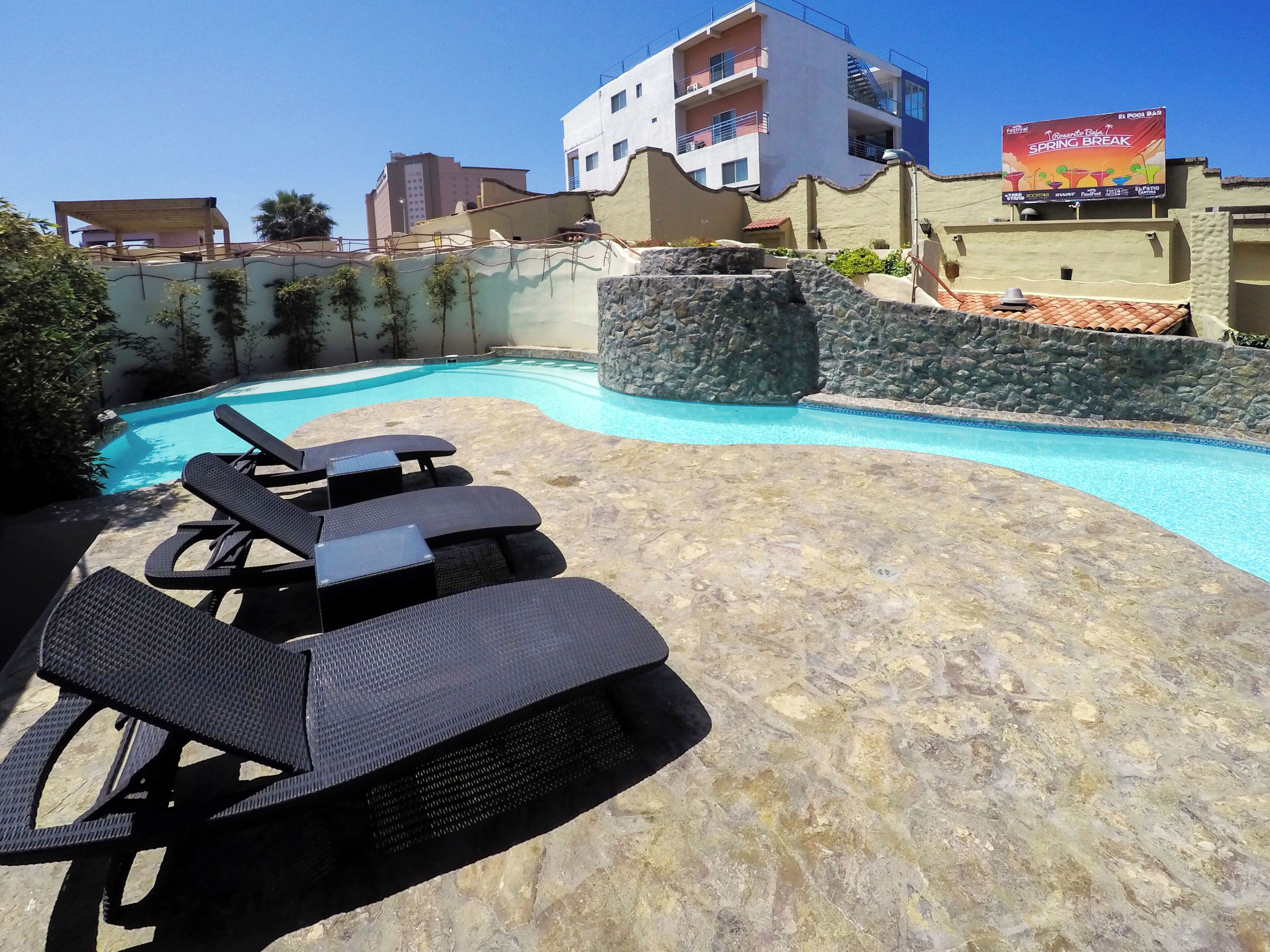 A swimming pool with lounge chairs, a stone wall, and a building with balconies in the background.