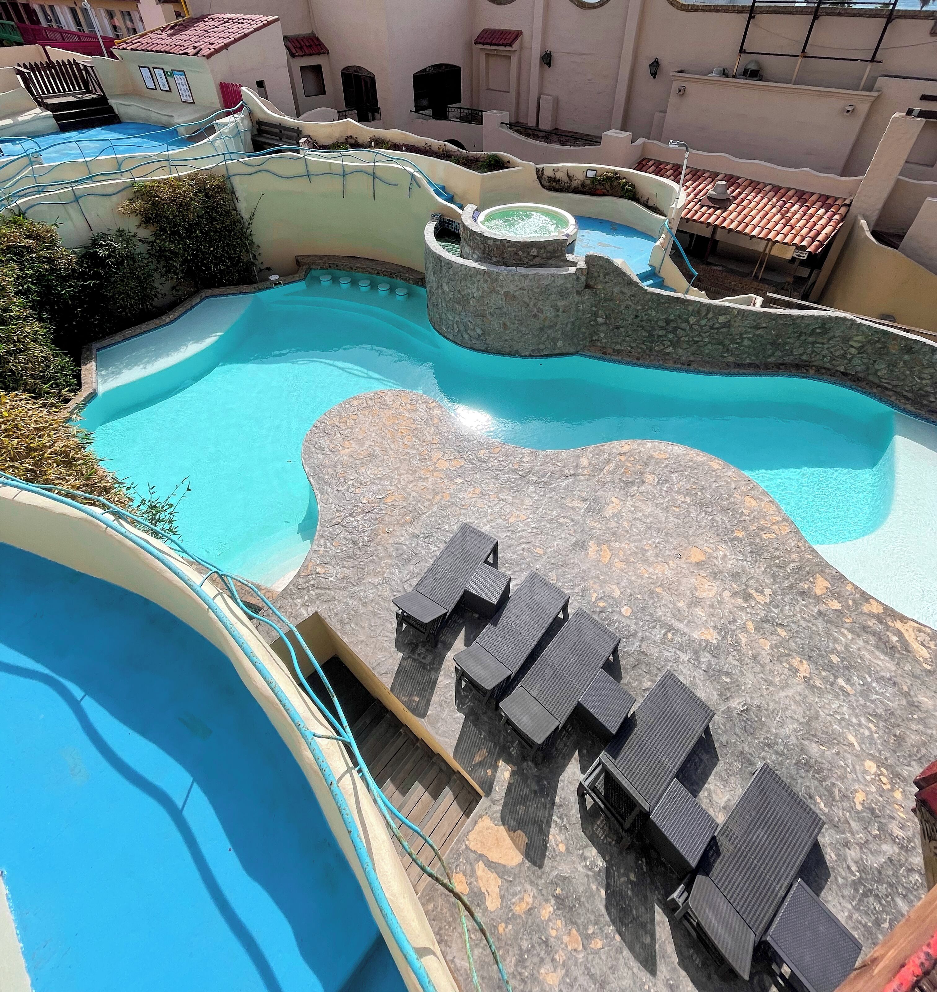 A swimming pool with a stone deck and lounge chairs, surrounded by buildings with red-tiled roofs.
