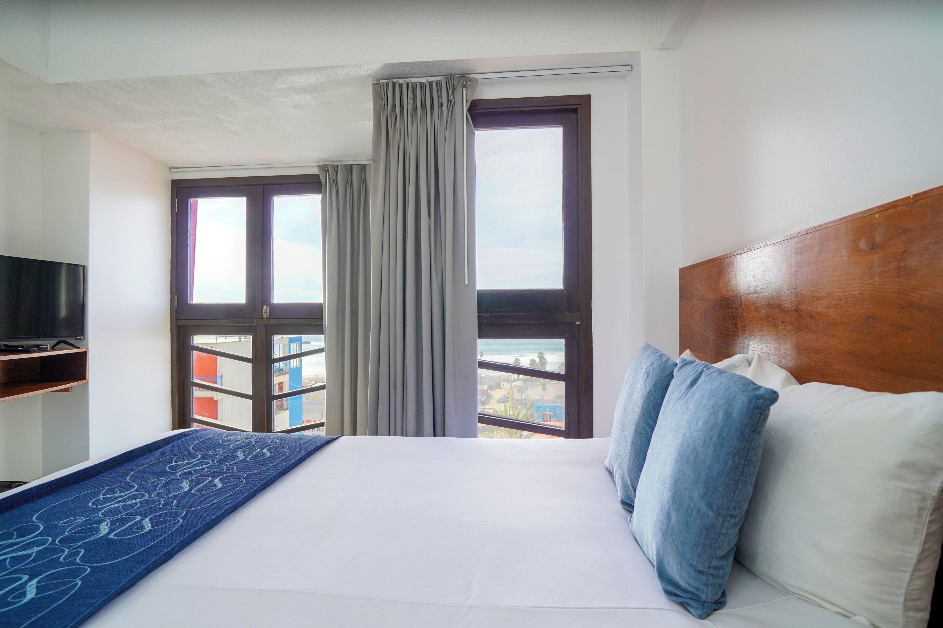 A hotel room with a bed, a TV, a window with curtains, and a view of buildings.