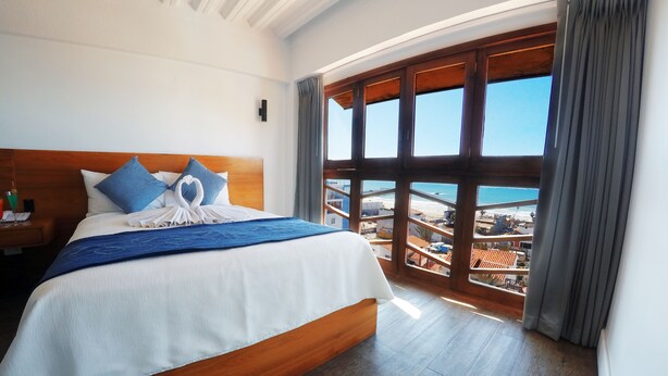 Ein Hotelzimmer mit einem großen Bett, einem Holzkopfende und Blick auf den Strand durch große Fenster.
