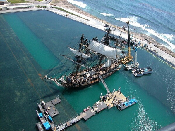 Luftaufnahme eines Schiffes, das an einem Steg liegt, mit einem großen Frachtschiff, kleineren Booten und einer Mole, die sich ins Wasser erstreckt.