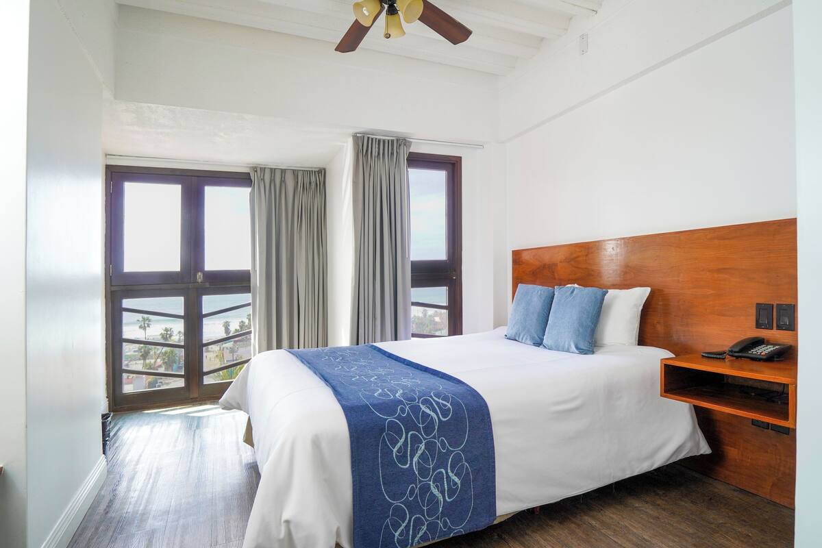 Habitación de hotel con cama, cabecera de madera, mesita de noche con teléfono y ventanas amplias con cortinas.