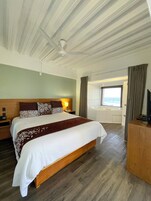 A hotel room with a large bed, a wooden headboard, a bedside table with a lamp, a TV, and a view of a pool and sky through a window.