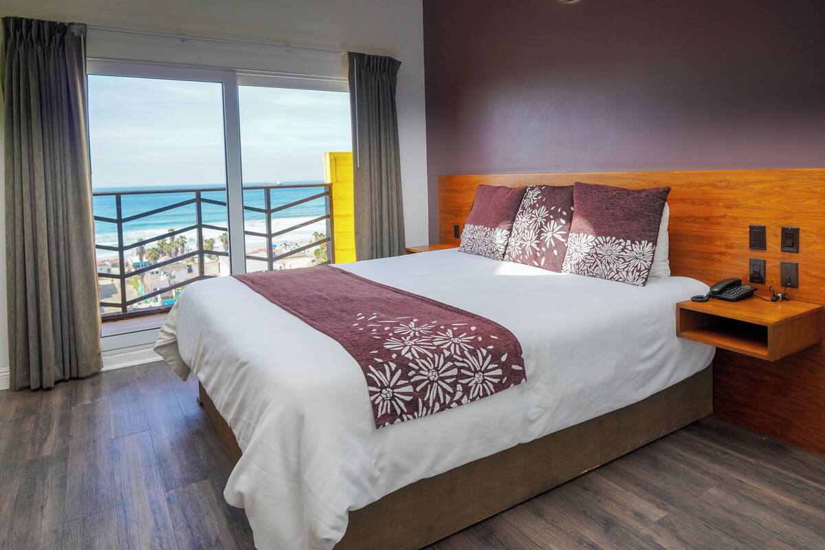 Una cama grande con cabecera de madera, una mesita de noche con un teléfono y control remoto, y vista al océano desde un balcón.