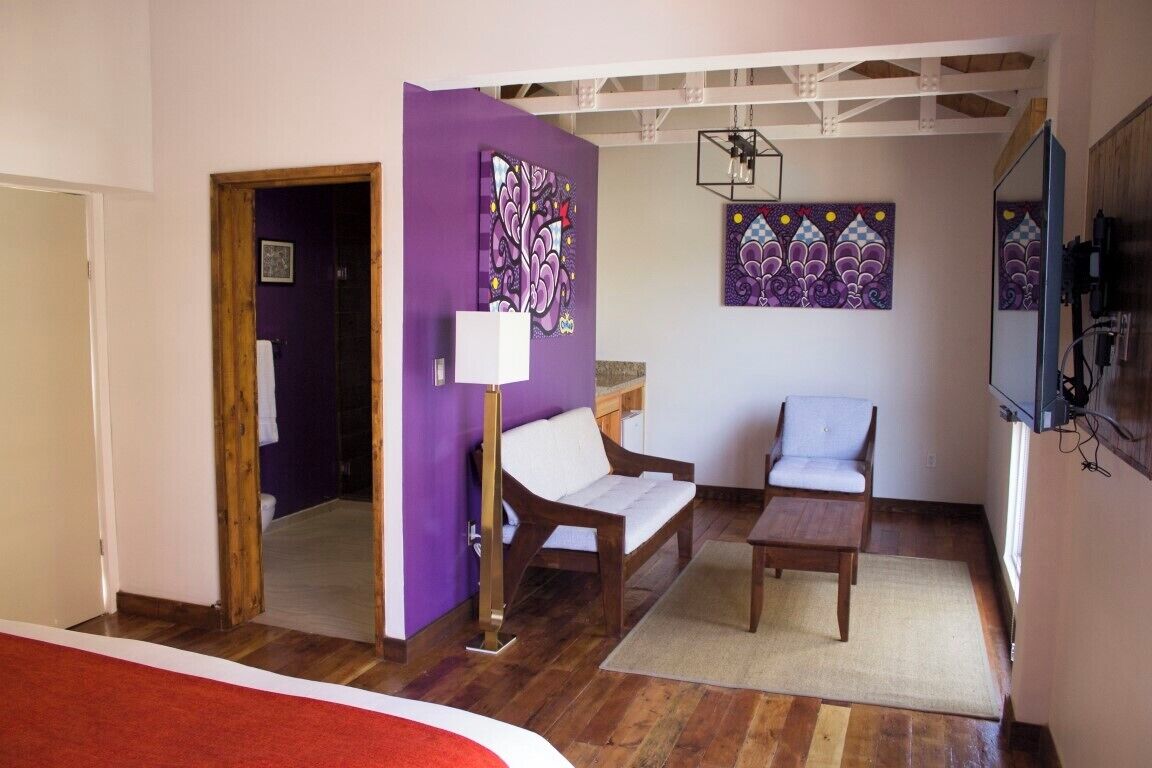 A room with a purple wall, a wooden floor, a bed, a white sofa, a wooden table, and two chairs.