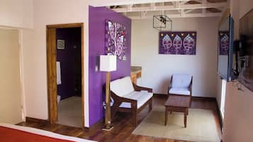 A room with a purple wall, a wooden floor, a bed, a white sofa, a wooden table, and two chairs.