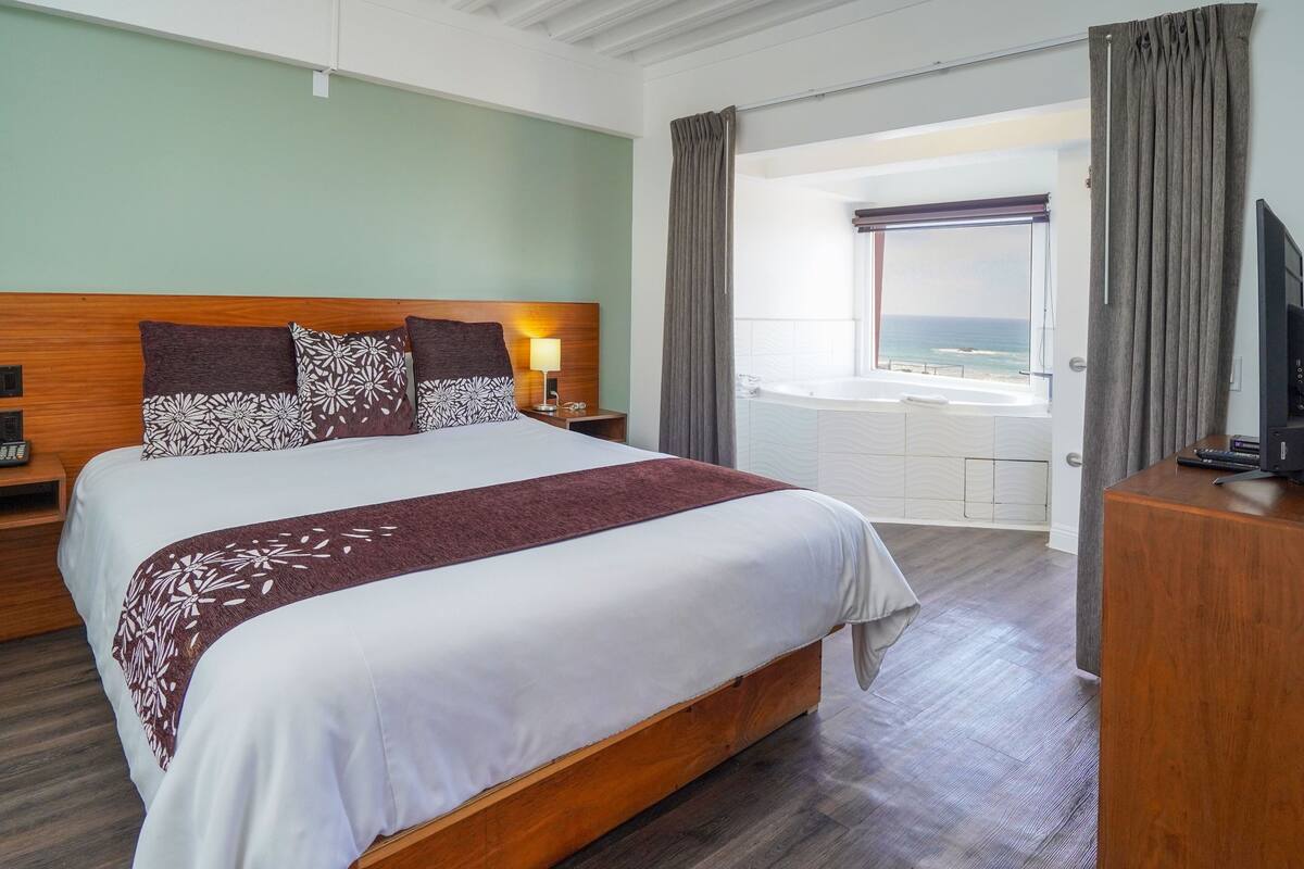 Habitación de hotel con una cama grande, televisión, mesita de noche, escritorio y vista al océano.