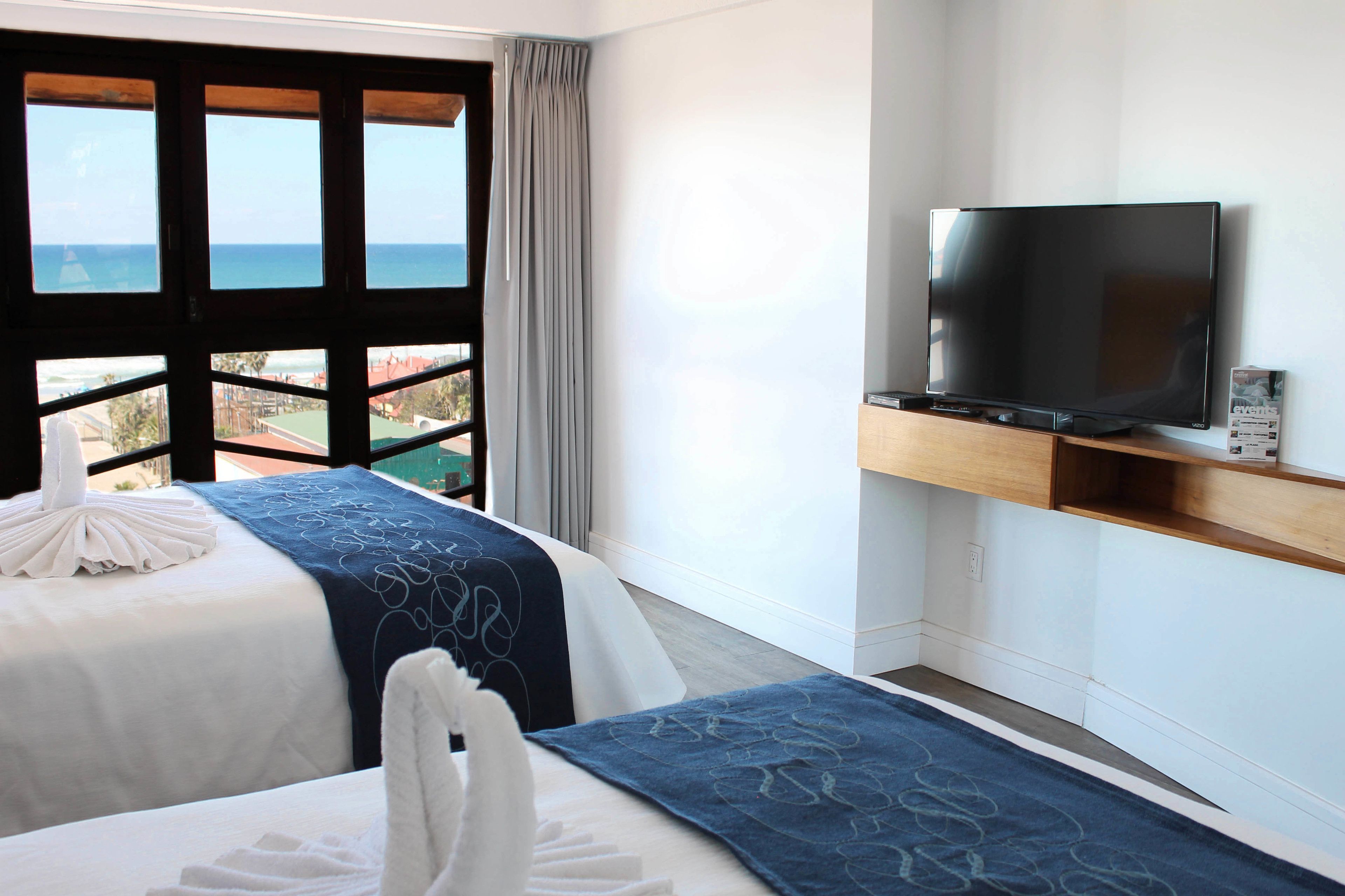 Une chambre d’hôtel avec deux lits, un téléviseur posé sur une étagère en bois et une vue sur l’océan grâce à de grandes fenêtres.