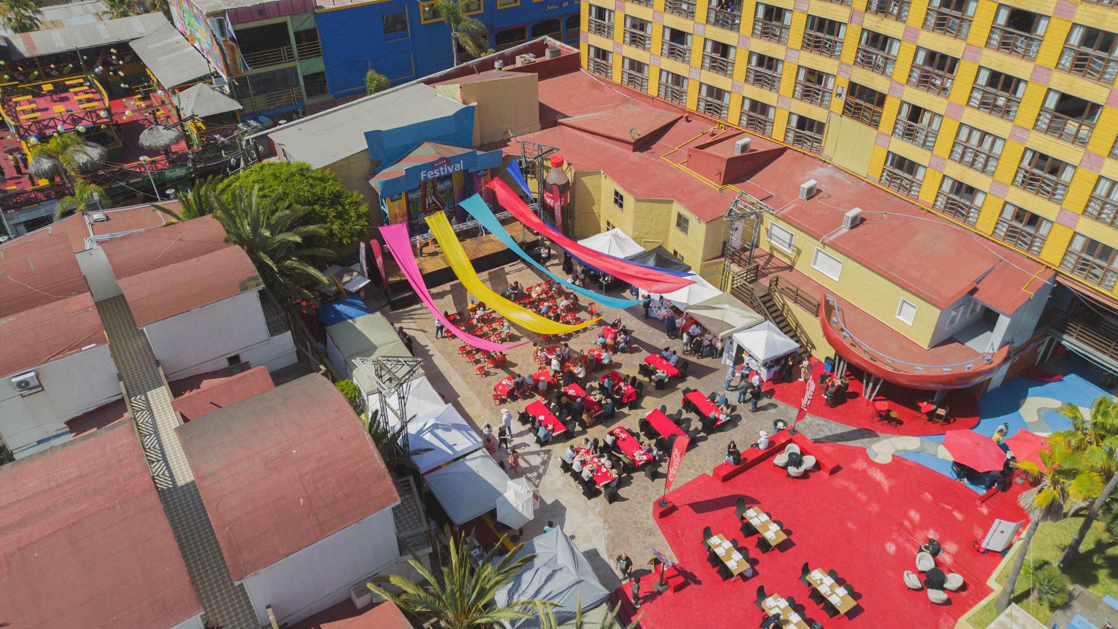 Un événement en plein air avec des tables et des chaises, une scène ornée d’une banderole Coca-Cola et un bâtiment à la façade jaune.