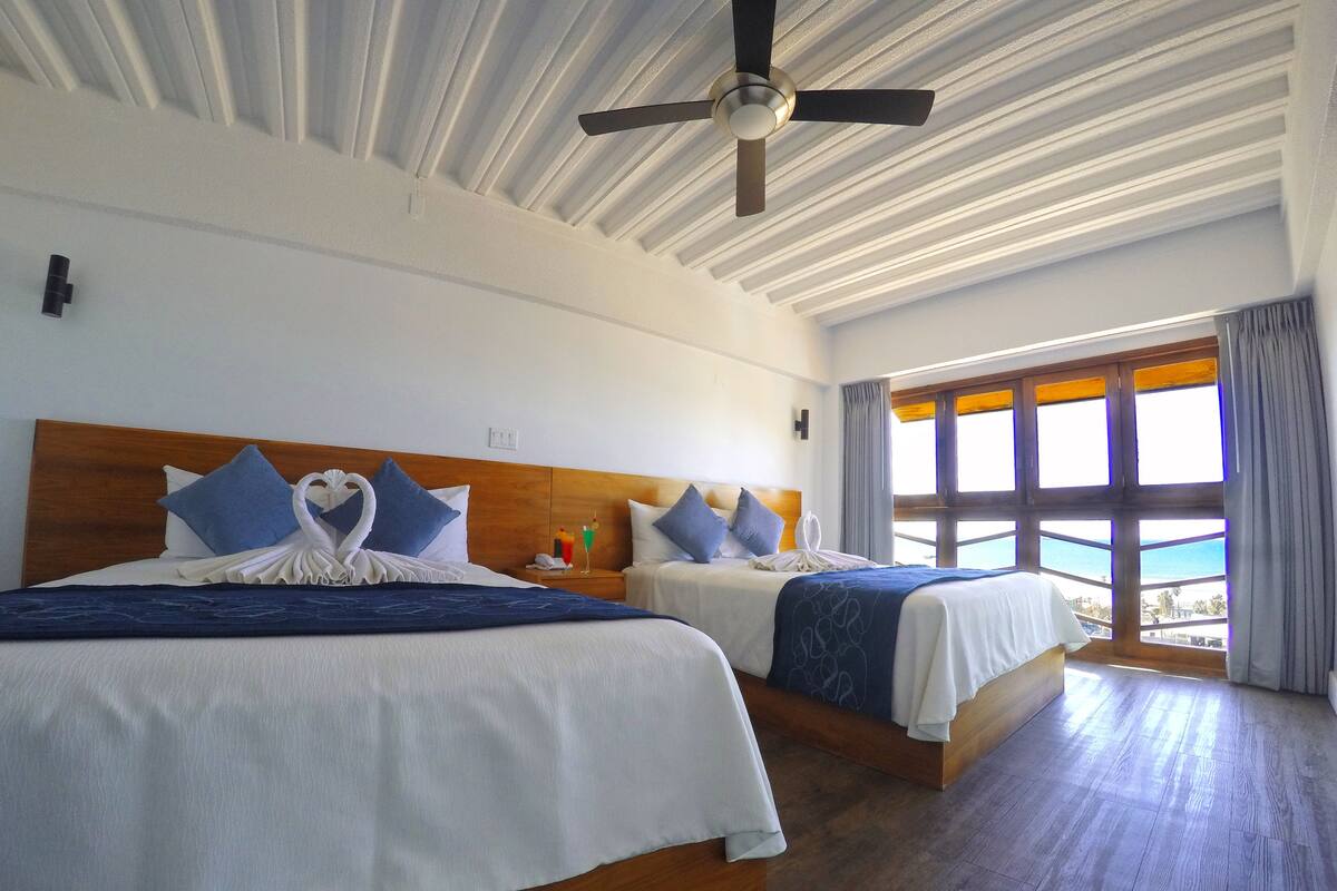 Habitación de hotel con dos camas, ventilador de techo y un ventanal con cortinas.
