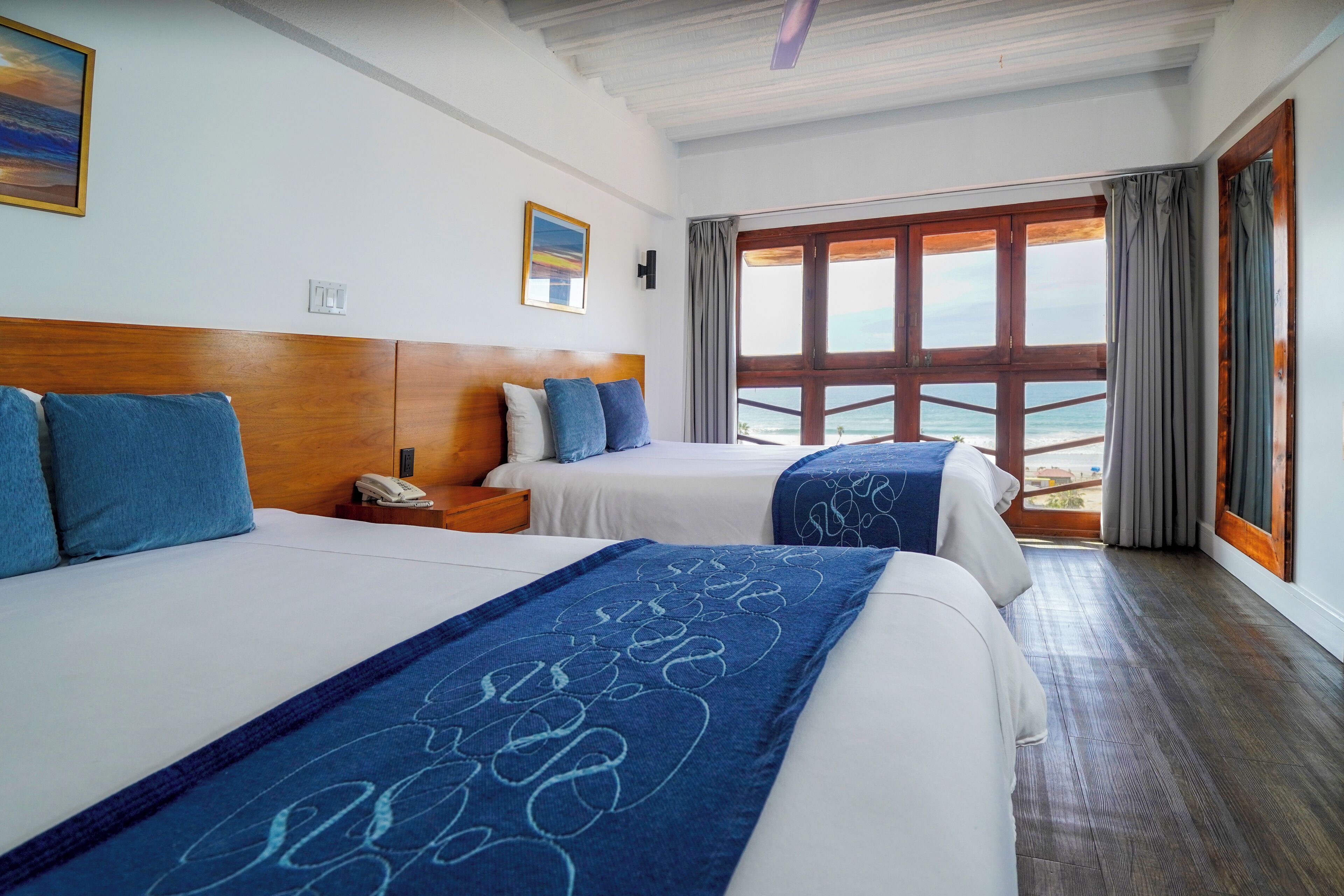 Habitación de hotel con dos camas, cabecero de madera, mesita de noche, teléfono y vista al mar a través de ventanas amplias.