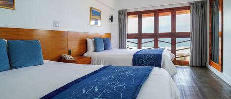 Habitación de hotel con dos camas, cabecero de madera, mesita de noche, teléfono y vista al mar a través de ventanas amplias.
