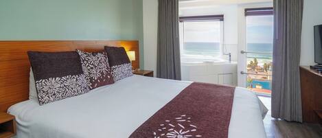 A hotel room with a large bed, a wooden headboard, a TV, and a balcony with a view of the ocean.