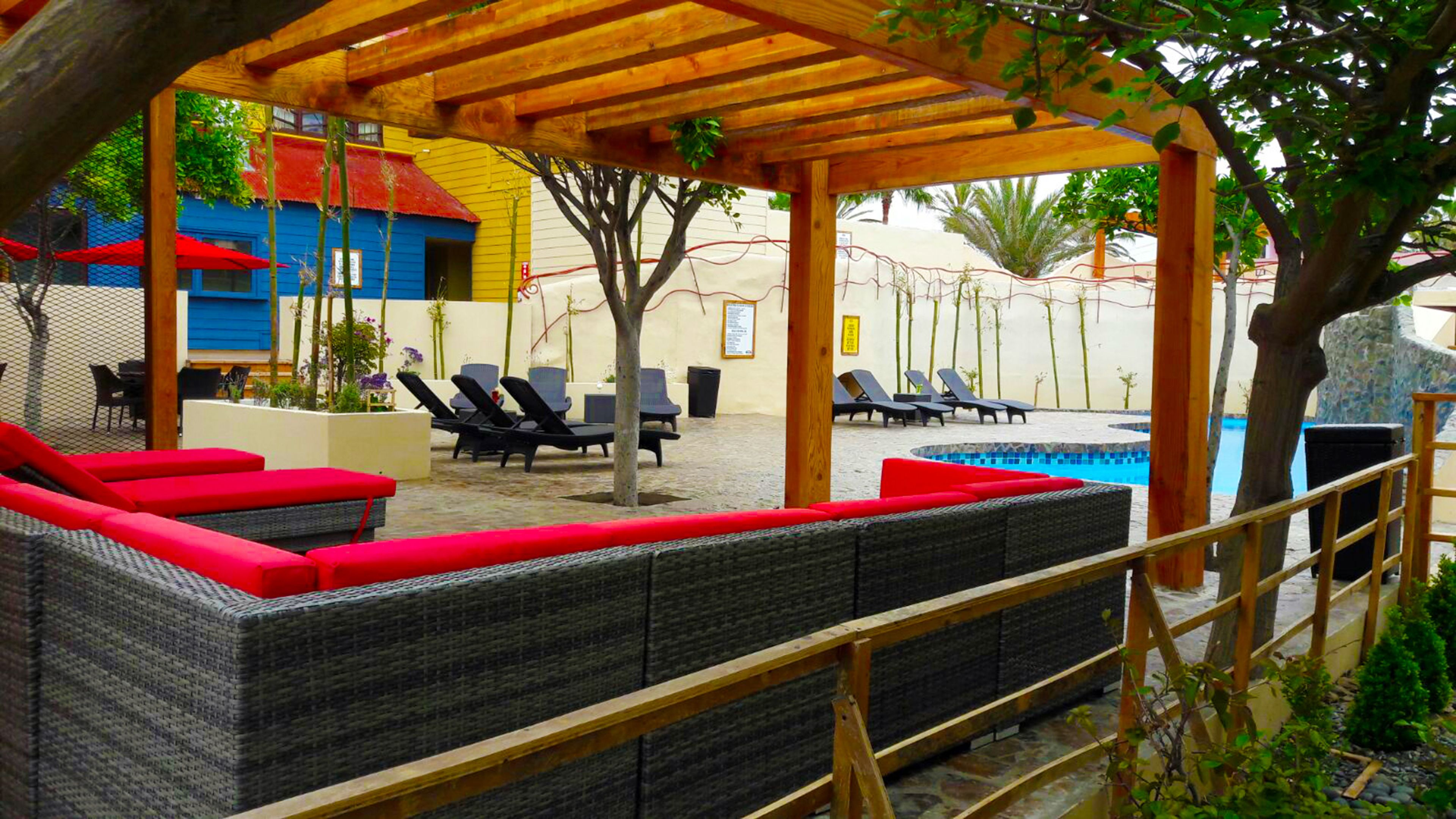 A covered outdoor seating area with wicker furniture and red cushions, overlooking a pool and a building with colorful facades.