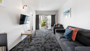 A modern living room with a flat-screen TV, a sofa with cushions, a coffee table, and a large window with curtains. - Lake Lodge Motel (Rotorua)