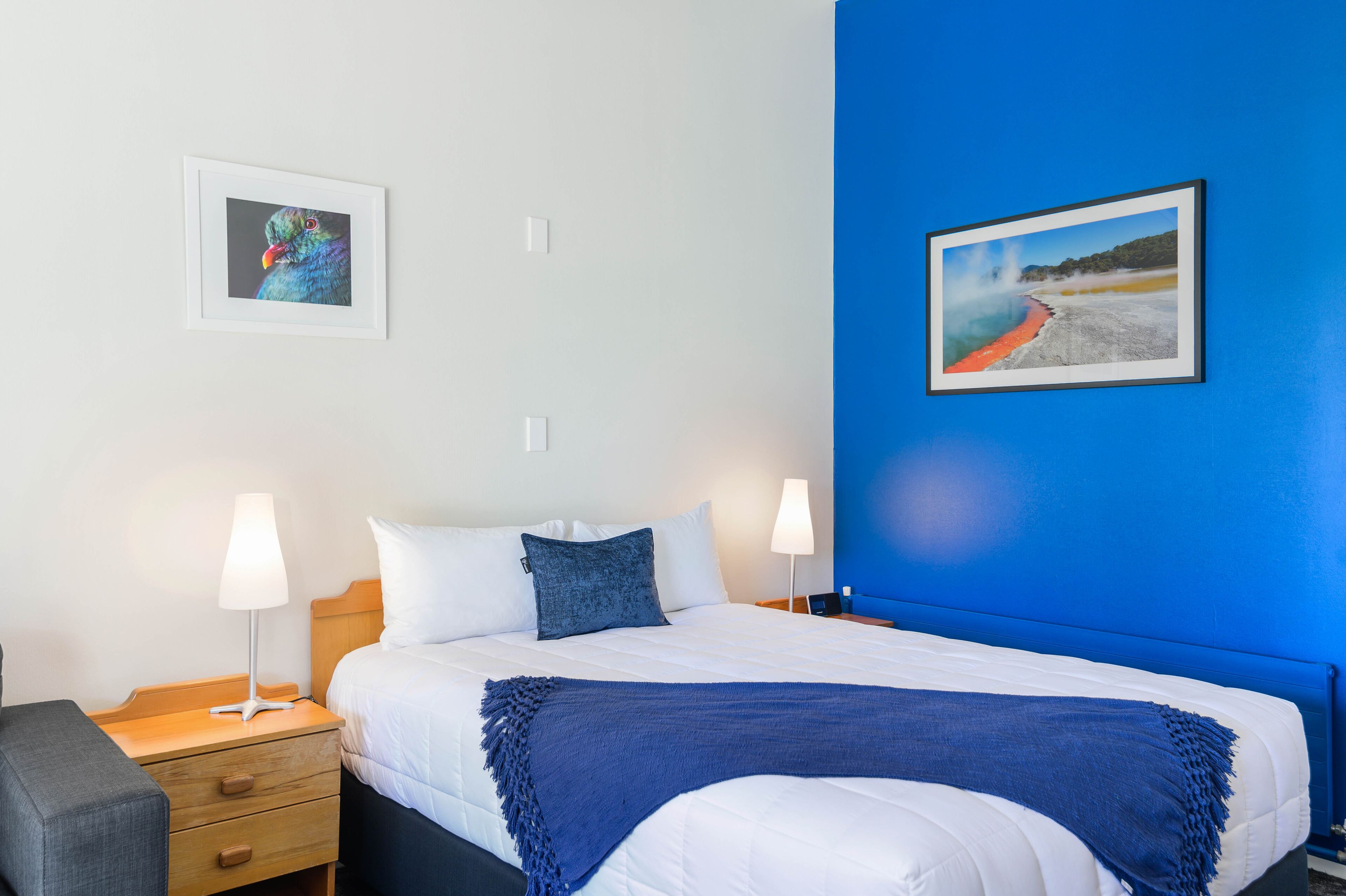 A hotel room with a bed, two bedside tables, a lamp, a chair, and two framed pictures on the wall.