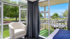 A room with a large window, a white armchair, a bed with a blue blanket, and a view of a parking area and trees. - Lake Lodge Motel (Rotorua)