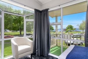 A room with a large window, a white armchair, a bed with a blue blanket, and a view of a parking area and trees.