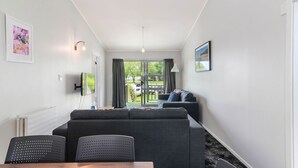 A modern living room with a grey sofa, a dining table with chairs, a flat-screen TV mounted on the wall, and a view of the outdoors through a glass door. - Lake Lodge Motel (Rotorua)