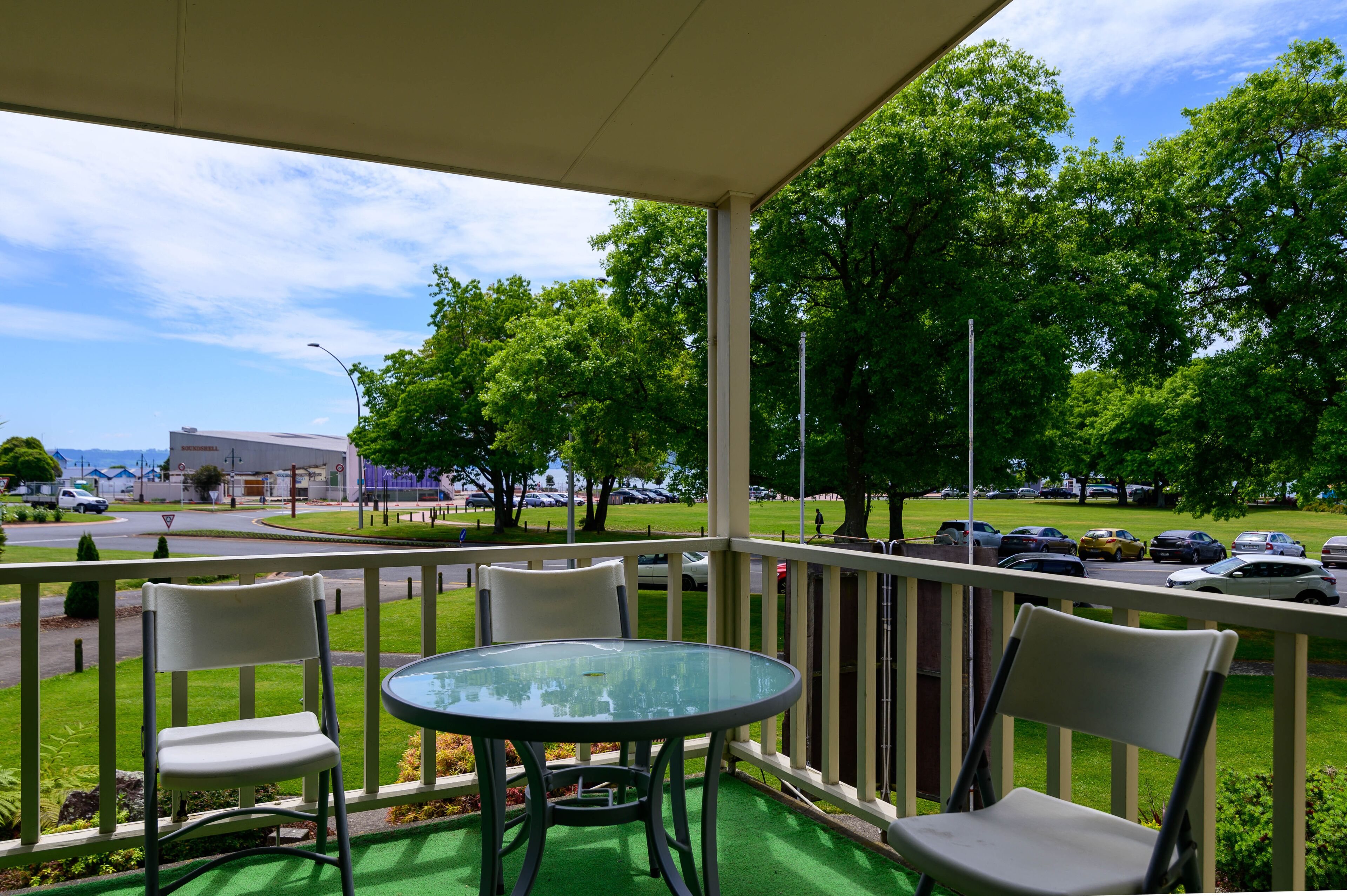 A covered outdoor seating area with a glass table and chairs, overlooking a parking lot and green space with trees.