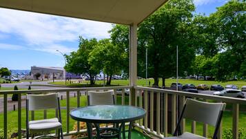A covered outdoor seating area with a glass table and chairs, overlooking a parking lot and green space with trees.
