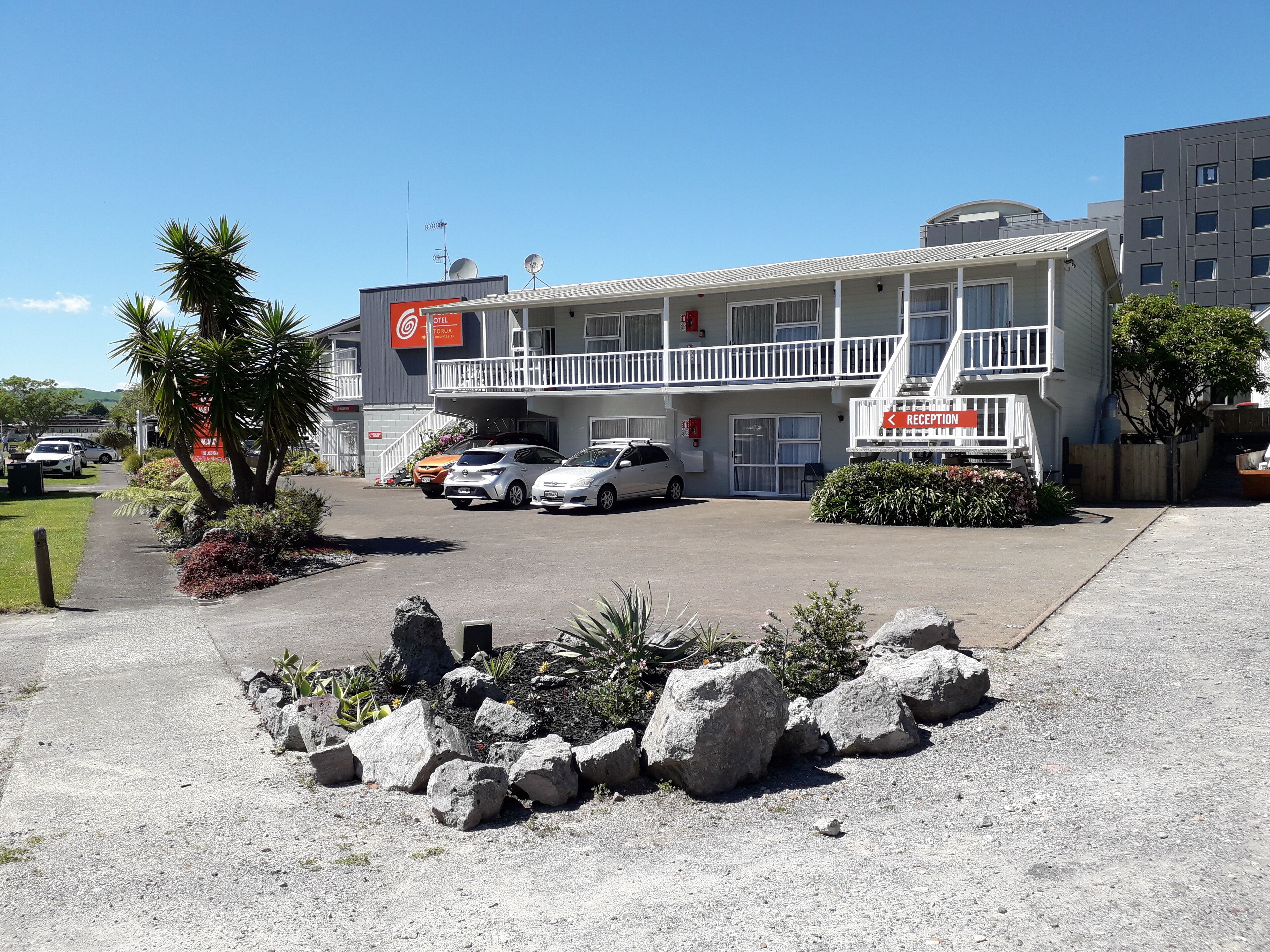 Um motel com área de recepção, várias vagas de estacionamento e um jardim frontal ajardinado com pedras e plantas.