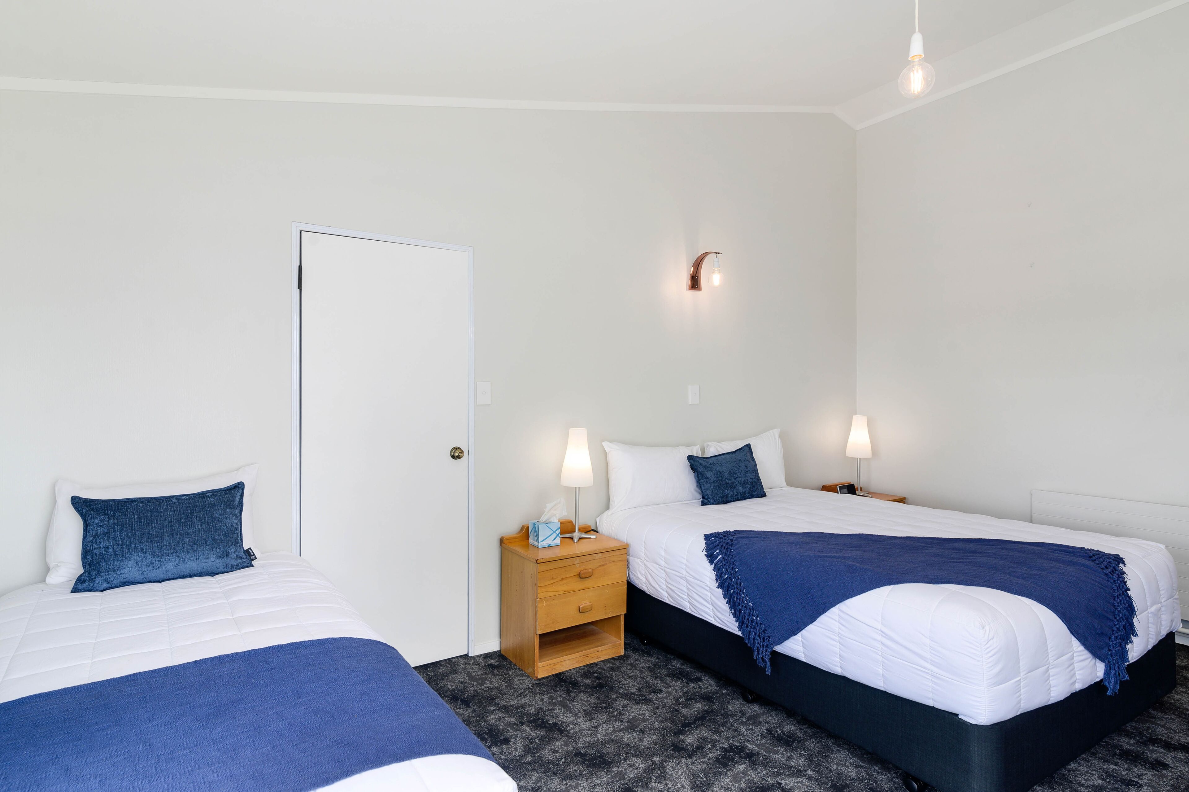 A bedroom with two beds, a nightstand, a door, and a wall-mounted light fixture.